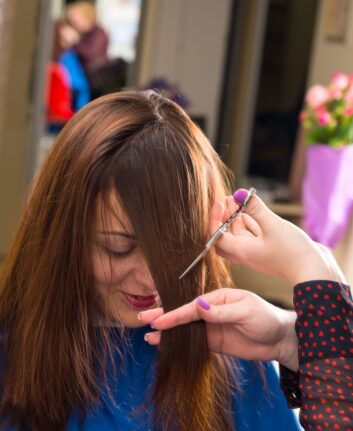 Close Up of Blond Stylist Cutting Hair of Young Brunette Client - Hair Dresser Cutting Angled Bangs into Hair of Young Female Client in Salon